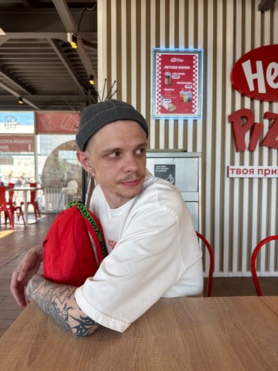 A man with a grey beanie and a white t-shirt sits at a wooden table in what appears to be a fast-food restaurant. He has tattoos on his arm and a nose ring. He is looking towards the camera. A red bag with green straps rests on his lap. Behind him, there is a wall with horizontal stripes and red lettering that reads "He" and "твоя при". There is also a menu displayed on the wall. The overall setting suggests a casual dining environment. Visible text in the image includes "ЛЕТНЕЕ МЕНЮ", "ЛИМОНАДЫ", "СПАСИБО ЧТО УБРАЛИ ЗА СОБОЙ", and "VIOBON KACO". The lighting suggests it is daytime.
