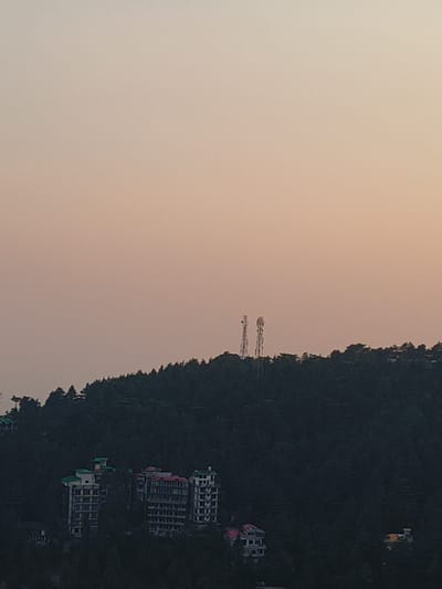 The image depicts a landscape scene at dusk with a view of hills covered in dense evergreen trees. Two tall cell towers are visible on the crest of a hill in the distance. In the foreground, several multi-story buildings with flat roofs are nestled among the trees, some featuring white and green facades, while others have red and white detailing. The sky is a gradient of soft orange and pale yellow, indicative of the fading light. The overall impression is that of a hillside community in a forested area.
