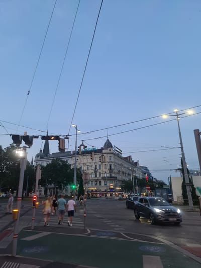 This is a vertical, eye-level shot taken from a street perspective in Vienna, Austria. The image appears to have been taken in the late afternoon or early evening, indicated by the soft, diffused light and the clear, pale blue sky. The presence of streetlights and traffic signals, some of which are illuminated, further suggests this time of day.
The scene is a busy urban intersection with a prominent building on the right side of the frame. This building features classical architecture with a domed structure and ornate detailing, suggesting it might be a historic or significant landmark. Several traffic lights are visible, with one displaying a yellow light, and a car, a dark SUV, is turning at the intersection. Pedestrians are crossing the street on a marked crosswalk. Some of the pedestrians are walking in groups, and their attire is casual, consistent with a warm evening.
The foreground is dominated by overhead wires, typical of a city with extensive public transportation, likely trams or trolleybuses. There are also lampposts and traffic signal poles. On the right side of the crosswalk, there's a green lane marked with bicycle symbols, indicating a bike path.
A few discernible text elements can be seen. On the building, "AUSTRIA" and "HOTEL" are partially visible in stylized lettering. There is also a sign that reads "CAFÉ". The text on the license plate of the car is illegible due to the image quality and motion blur.
The overall mood of the image is one of urban life continuing as day transitions to evening, with a sense of movement and activity. The clear sky and lack of rain suggest pleasant weather.