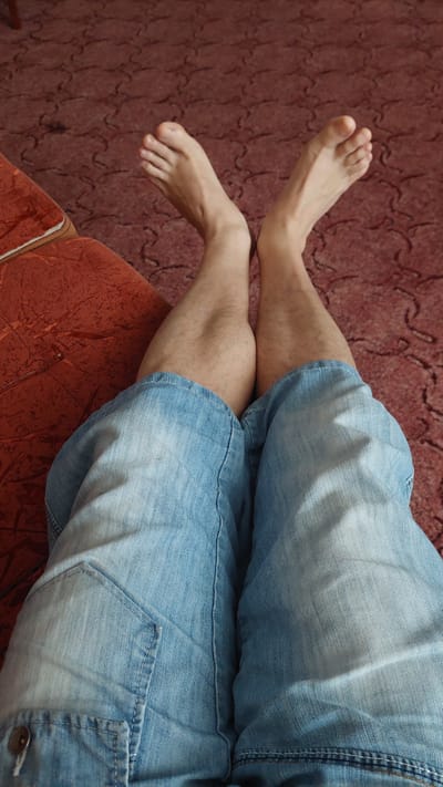 The image shows a first-person perspective of a person's legs and feet, resting on a red patterned carpet. The person is wearing light blue denim shorts. Their legs are crossed, with the feet positioned upwards. The scene appears to be indoors, in what could be a living room or bedroom, given the carpet and the hint of an upholstered piece of furniture on the left side of the frame. There is no indication of the specific location within Chișinău, Moldova, as the image does not contain any recognizable landmarks or contextual clues.