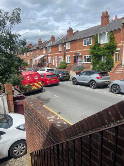 A street scene in the city of Reading, United Kingdom, on a cloudy day. The image shows a row of red-brick terraced houses on the right side of the street, with a tree and more houses on the left. Several cars are parked on the street, including a red Royal Mail van, a red hatchback, a grey Volvo SUV, a grey sedan, a white hatchback, and a black hatchback. A "FOR SALE" sign is visible on the front lawn of one of the houses. The street is paved with asphalt and has a yellow line painted on the side. There are dustbins and some overgrown ivy on the houses. The foreground shows a brick wall and metal railings.