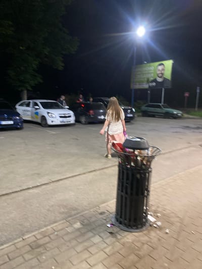 The image depicts a nighttime scene in Poltava, Ukraine, possibly at a gas station or rest stop, with a bright overhead lamp illuminating the area. In the foreground, a black, cylindrical trash can overflows with garbage, with debris scattered around its base on a paved walkway. A young woman with long brown hair, wearing a white top and a pink skirt, walks past the trash can, carrying a pink bag. Behind her, several cars are parked: a dark blue sedan on the left, a white hatchback with the "Uklon" logo and a Ukrainian flag sticker, and a dark hatchback further back. In the background, a large billboard features a portrait of a smiling man. The environment appears to be an open, paved area adjacent to a road, with trees visible on the left.