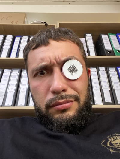 The image shows a close-up selfie of a man with a beard. He is wearing a black t-shirt and has a white circular object with a QR code on it placed over his left eye. He is looking directly at the camera with a somewhat surprised expression. Behind him, there are shelves filled with binders. The man is positioned in an office environment. The location is identified as Luzhitsy, Russia, suggesting an interior setting within a building in that city.