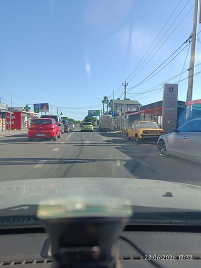 The image shows a street scene in Lymanka, Ukraine, on a sunny day. The perspective is from inside a vehicle, looking forward. Several cars are on the road, including a red Hyundai, a yellow car, and a silver car. Buildings line the street, with some storefronts visible. Power lines and streetlights are present. A traffic light is visible in the distance. The timestamp on the image reads "22.06.2026 17:32".