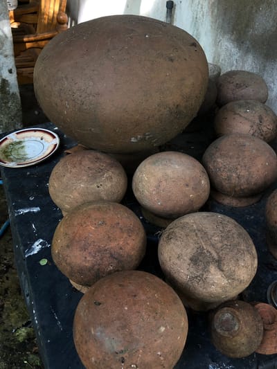 A collection of clay pots, varying in size and condition, are arranged on a dark surface. The largest pot, situated at the back and center, has a weathered, globular shape with visible soil and debris. Surrounding it are numerous smaller, spherical pots, some stacked or clustered together. A small, ornate plate with a red and white rim and some green residue is visible on the left. The background consists of a textured, light-colored wall and hints of wooden furniture. The scene suggests an outdoor storage or display area, possibly in Kabalana, Sri Lanka, given the implied context. The lighting is diffused, casting subtle shadows.