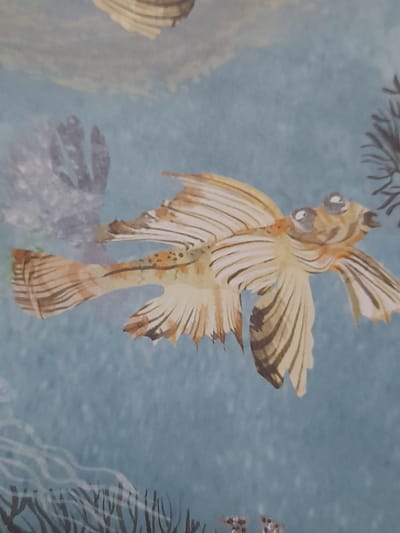 The image is a close-up shot of a decorative pattern on a blue background. The pattern features a stylized depiction of a yellow and brown fish with large, bulging eyes. The fish has prominent fins and tail, with dark stripes accentuating its form. The background appears to be a textured wallpaper or fabric, with hints of other abstract shapes and elements that might represent underwater flora or other marine life. There are no people or identifiable landmarks from Baan Mae Nam, Thailand, in this image.