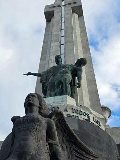 The image shows a low-angle view of the Monument to the Fallen (Monumento a los Caídos) in Santa Cruz de Tenerife, Spain. The monument features a tall, stone tower with a cross at the top. In the foreground, there are several bronze sculptures. The most prominent are two figures at the base of the tower: a seated female figure holding a deceased male figure in her arms. Below these figures, and closer to the camera, is a large, dark stone sculpture of a winged figure, likely an angel, looking upwards. The inscription "A TODOS POR LOS" is visible on the base of the monument in green lettering. The sky is partly cloudy with a mix of blue sky and white clouds. A pigeon is perched on the wall in front of the inscription.