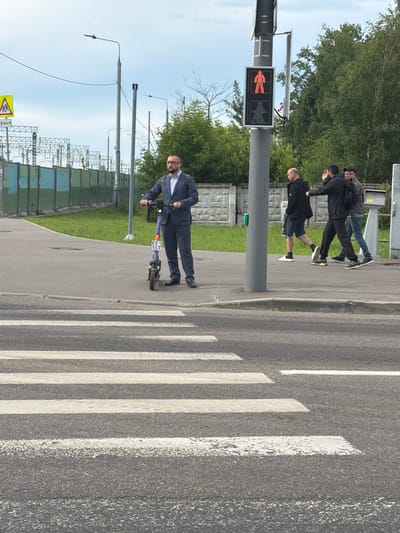 In Shcherbinka, Russia, a man in a dark suit stands on a paved sidewalk next to a purple electric scooter. He is facing left, with his hands on the scooter's handlebars. A red pedestrian signal displays a walking person, indicating it is safe to cross. Behind him, three men walk on the sidewalk, two in dark clothing and one in a denim jacket. The background features trees, a fence, and electrical infrastructure, suggesting an urban or suburban setting. A crosswalk is visible in the foreground.