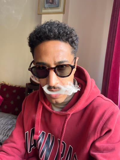 A man with dark curly hair and brown skin is wearing tortoise-shell framed sunglasses and a red hooded sweatshirt. He is exhaling a plume of white smoke from his mouth. The setting appears to be indoors, with a red velvet headboard visible at the top and a patterned cushion partially visible at the bottom. There is no evidence to determine if the location is Tanger, Morocco.