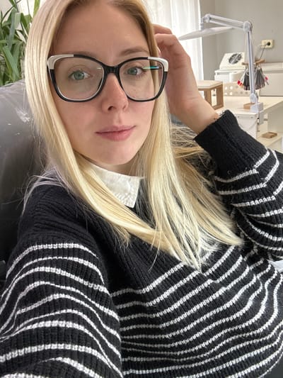 A blonde female, appearing to be a young adult, is centered in a medium shot, looking directly at the camera with a neutral expression. She has fair skin, green eyes, and wears black-framed glasses with white accents on the upper outer corners. Her long, straight blonde hair is loosely styled, with her left hand subtly touching the hair on the right side of her head. She is dressed in a black knitted sweater with multiple white horizontal stripes, worn over a white collared shirt. The indoor background suggests a professional setting, possibly a salon or studio. To the left, a dark object, likely a chair, and a portion of a green plant are visible. To the right, a white workstation features an articulated white desk lamp clamped to the surface, a square white device with a fan grille (consistent with a dust collector), and a fan-shaped holder displaying numerous colored nail polish swatches. A light-colored wooden stool or stand is also present on the desk. A power outlet with a plugged-in cord is visible on the light-colored wall above the desk, and a window with a sheer white curtain is partially seen in the upper right background. The specific location within the city of Voskresensk, Russia, is not discernible from the visual information presented in the image.