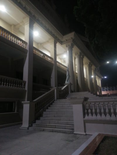The image depicts the exterior of a grand building at night, characterized by a classical architectural style. A wide, illuminated staircase dominates the foreground, leading up to a colonnade with tall, fluted columns supporting a decorative entablature. A statue of a figure stands to the left of the stairs, near a balcony with ornate railings. The building's facade features multiple levels with balconies and is illuminated by interior lights, casting a warm glow. Dense foliage, likely trees, is visible against the dark night sky on the right side of the frame. The ground in front of the stairs is paved. The overall impression is one of a stately and well-maintained public building, possibly a museum or government structure, located in a city with a temperate climate given the presence of lush trees. The specific location in Xuân Hương - Đà Lạt, Vietnam, suggests a possible cultural or historical landmark within the city.