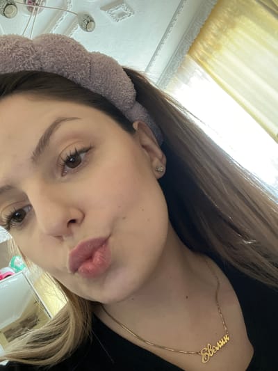 A close-up, angled selfie of a woman with long brown hair wearing a light purple fuzzy headband. She has brown eyes and is pursing her lips. A delicate gold necklace with a word in Cyrillic script rests on her neck. Her right ear is visible with a small flower-shaped earring. The background includes a yellow curtain and a white wall with decorative trim. The lighting is bright, possibly from a window. There is no information available to definitively confirm that this image was taken in Novi Pazar, Bulgaria.