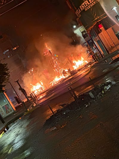 The image depicts a scene of widespread fire and destruction in what appears to be an urban environment at night, likely Alicante, Spain, based on the presence of a banner with text that appears to be Spanish. A large structure, possibly a temporary construction or monument, is engulfed in flames and smoke, with debris scattered on the wet street below. The street is reflective, indicating recent rain or water use, possibly from firefighting efforts.
In the foreground, the street surface is dark and wet, with puddles and patches of standing water reflecting the orange glow of the fire. Debris, including what looks like burnt materials and possibly trash, is scattered across the roadway. A yellow hose is visible snaking across the street, suggesting active firefighting or its aftermath. There are several poles and street furniture, some of which appear damaged.
In the mid-ground, the main subject is a large, multi-tiered structure that is almost entirely consumed by fire. Flames are erupting from various levels, and thick smoke billows upwards and outwards, obscuring parts of the surrounding buildings. The structure has a skeletal appearance, with exposed wooden or metal framework visible through the fire.
In the background, buildings line the street. One building has a large dark green banner with white lettering, which appears to read "ESTRELLA DE LEVANTE". There are windows visible in the buildings, and in one, several people are seen looking out, observing the scene. Another building has illuminated signage and what looks like a storefront. The overall lighting is dominated by the fire, casting an orange and red hue over the entire scene. The image conveys a sense of chaos and destruction.