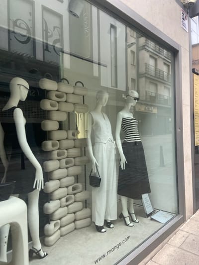 The image shows the storefront of a Mango clothing store located in Segovia, Spain, at the coordinates 40.946, -4.119. The storefront is a glass display case, and there are three mannequins inside. The mannequin on the left is wearing a black dress and black shoes. The mannequin in the center is wearing white pants, a white sleeveless top, a black purse, and black shoes. The mannequin on the right is wearing a black and white striped top, a black skirt, and black shoes. The mannequins are standing in front of a wall of white, rounded, stacked objects, and a white wall behind them. The exterior of the store is made of light-colored stone. The storefront is angled towards the viewer, and a sign with the text "www.mango.com" is visible in the bottom right corner of the image. There are buildings and streetlights visible in the background, through the storefront windows.