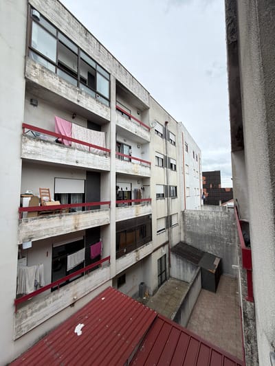 The image depicts a high-angle view into a narrow urban space between residential buildings in Bragança, Portugal, under an overcast sky. The foreground features a partial red corrugated metal roof.
To the left, a multi-story concrete apartment building exhibits weathered surfaces with peeling paint and exposed concrete. Its facade features several balconies with red metal railings.
* The uppermost visible balcony has a large glass window section above it and a pink fabric piece and a white, patterned fabric hanging over its railing.
* The middle balcony on the left displays a wooden rocking chair, a white box-like object, and a brown cardboard box. A white roller blind is partially lowered behind the railing.
* The lowest visible balcony on the left has white and pink laundry hanging over its railing and additional white laundry items suspended on a line further back.
Centered in the mid-ground, another multi-story building, lighter in color, is visible. It also features balconies with red railings, one of which has a small satellite dish attached. Laundry is visible hanging on multiple balconies of this building. A vertical drainpipe runs down its facade.
The ground level, mostly obscured by the foreground roof, reveals a concrete-paved courtyard area. Two small, dark-roofed structures, resembling sheds, are situated in this area. A dark-colored waste bin stands near the base of the center building's wall. A tall, textured concrete wall, part of an adjacent building, frames the right side of the image, showing similar signs of wear. Beyond the courtyard, in the background, another building with a dark, possibly tiled roof is partially visible. No individuals are present in the frame.