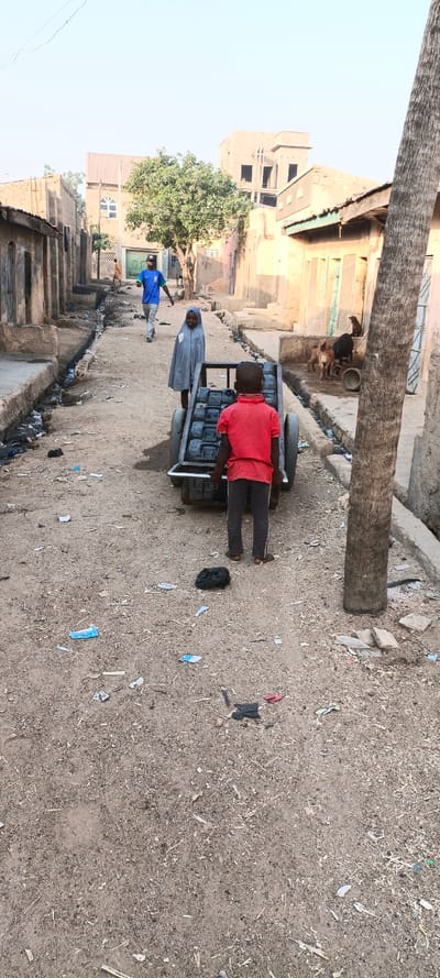 A vertical shot captures a dusty, unpaved street scene in Kaura Goje, Nigeria, under a bright, clear sky, suggesting either morning or late afternoon. In the foreground, a young boy in a red short-sleeved shirt and dark pants, barefoot, stands with his back to the camera, appearing to push a metal handcart. The cart is heavily loaded with multiple dark, rectangular containers, likely jerrycans used for transporting water.
To the left of the cart, a young girl wearing a grey hijab and a loose-fitting robe stands, looking towards the camera with a neutral expression. Further down the street, a man in a blue t-shirt and light-colored pants walks towards the viewer. Another person in lighter clothing is visible in the distance on the left side of the street.
The street is lined with various buildings, predominantly single or two-story structures, some appearing unfinished or in a state of disrepair. A leafy green tree stands in the mid-ground on the left, providing a focal point amidst the buildings. On the right side of the frame, the thick trunk of a palm tree partially obscures a view of additional buildings and several goats, possibly with a dog, gathered near a drainage ditch that runs parallel to the street.
The ground throughout the image is covered in sand and dirt, scattered with debris, including plastic wrappers, paper, and wood chips, indicating unpaved conditions and visible litter. The drainage ditches flanking the street also contain accumulated trash. The scene portrays daily life, highlighting activities like water transport in a residential area with a visible lack of modern infrastructure. No discernible text is visible.
