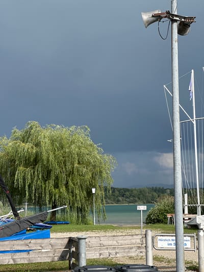 This image depicts a lakeside scene in Chiemsee, Germany, under a dark, cloudy sky. In the foreground, a weathered wooden fence runs horizontally across the frame. To the right of the fence, a metal gate with a sign reads "Herzlich Willkommen im S" (Welcome to the S...). Behind the fence, several sailboats or dinghies are covered with blue tarps, and a large weeping willow tree dominates the mid-ground. A lamppost stands near the tree, and beyond that, a clear turquoise lake stretches towards a tree-lined shore and distant hills. On the right side of the image, a tall metal pole supports a large loudspeaker and a smaller siren or bell. The sky is overcast with dark storm clouds gathering.
