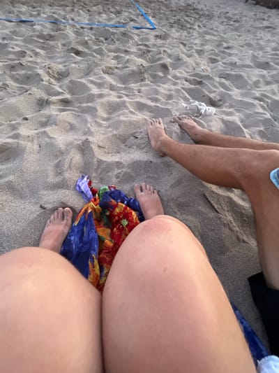 The image shows two people's legs and feet on a sandy beach. A colorful sarong or towel is draped over the sand between them. In the background, a blue net or rope is visible, possibly part of a beach volleyball court. The sand is rippled and appears to be a mix of light and dark grains. The lighting suggests it is either early morning or late afternoon. The scene is located on a beach in Miljevci, Montenegro.