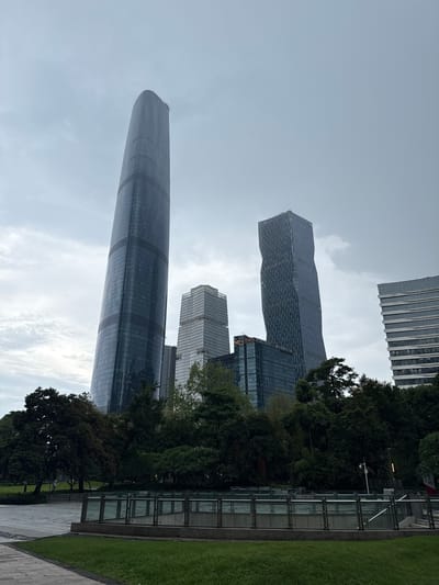 The image shows a panoramic view of modern skyscrapers in the Tianhe District of Guangzhou, China, under an overcast sky. The foreground features a well-maintained green lawn and a paved walkway. In the mid-ground, lush green trees form a dense line, partially obscuring lower levels of the buildings. A glass-paneled barrier with metal railings runs across the mid-ground, suggesting a park or public space.

Dominating the scene are several tall, contemporary buildings. The most prominent is a massive, sleek, dark-glass skyscraper with a distinctive curved, tapering form, leaning slightly to the left. To its right, another skyscraper stands, characterized by its angular, twisted design with a dark facade. Further to the right, a rectangular, light-colored building with numerous horizontal window bands is visible. Other buildings, including one with the "ASCOTT" logo, are also present, contributing to the urban skyline. The overall impression is one of a bustling, modern city district characterized by impressive architecture. There are no people visible in the image.