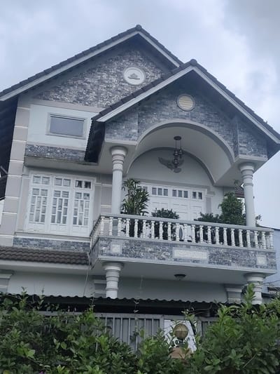 A light gray, three-story house with a dark gray tiled roof is pictured in Lâm Viên - Đà Lạt, Vietnam. The house features a prominent arched entrance on the second floor, supported by two fluted columns and leading to a white double door. A balcony with a decorative white railing runs across the second floor, adorned with grey stone accents. The windows on the second floor are white with intricate pane designs, while the third floor has a smaller, rectangular window. The exterior walls are a mix of light gray paint and grey stone tiles, particularly around the gables and balcony. A chandelier hangs from the ceiling of the arched entrance, and a bird relief is mounted on the back wall of the arch. Lush green foliage and a metal fence are visible in the foreground. The sky is overcast.