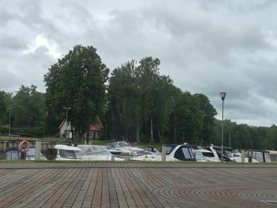 The image shows a marina with several boats docked along a concrete pier. The boats are of various sizes and types, with some covered by tarps. In the background, there are trees and a building, which appears to be a house. The sky is overcast, suggesting cloudy weather. The foreground is a paved walkway with a patterned brick design. A lamppost is visible to the right of the frame. The location is the Šyša River marina in Šilutė, Lithuania. There are no people visible in the image. The scene is peaceful and depicts a typical day at a marina.