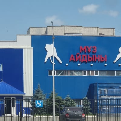 The image shows the exterior of a blue building with signage and a white silhouette of a person in a dynamic pose, possibly jumping or dancing. Red lettering on the building reads "МУЗ АЙДЫНЫ" which translates to "Music Aisyn" or "Music Echoes." The building appears to be a cultural center or event space, given the imagery.
A handicap parking sign is visible on the right side of the image, indicating accessibility. The scene suggests daytime with clear skies and sunlight casting shadows, typical of good weather. The building is modern with a mix of blue and white sections, and windows are visible. A dark-colored car is parked near a black metal fence in front of the building. Several evergreen trees are planted near the building. In the distance, small dark shapes in the sky might be birds or drones.
The overall impression is a bright, modern public building in a location that experiences sunny weather. The stylized silhouette and bold signage suggest a place that hosts lively events or performances. The context provided places this scene in Aktobe, Kazakhstan.