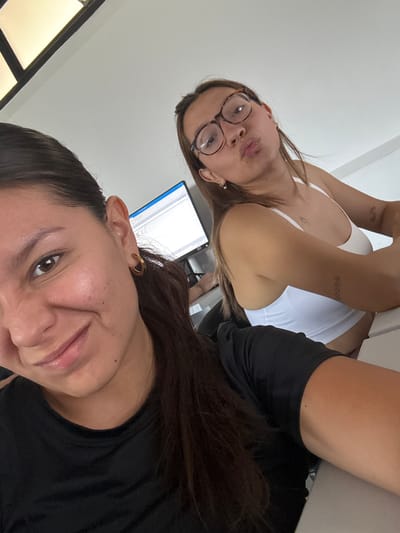 This media file features two women indoors, likely in San Andrés Cholula, Mexico, captured in a casual, low-angle selfie. The image is rotated counter-clockwise.
In the foreground, partially filling the lower right portion of the rotated image, is the face of one woman. She has dark hair, dark eyes, and a subtle smile, with a visible gold hoop earring on her ear. Her dark top is visible at her shoulder and neck.
Behind her, occupying the upper left of the rotated frame, is the second woman. She has light brown hair and is wearing tortoiseshell-framed glasses. Her lips are playfully puckered in a "duck face" expression. She wears a white tank top, revealing several small tattoos on her arms, including what appears to be a butterfly on one arm and a linear pattern of small stars on the other.
The setting is an indoor environment with light-colored walls or ceiling, suggesting a bright room. A computer monitor displaying what looks like a document or code with a blue scroll bar is partially visible in the background, hinting at an office or home office setup. The overall mood is relaxed and friendly. No explicit text is clearly legible in the image, apart from the unreadable content on the screen. The time of day is not discernible, as the lighting appears to be artificial or from an unseen window.