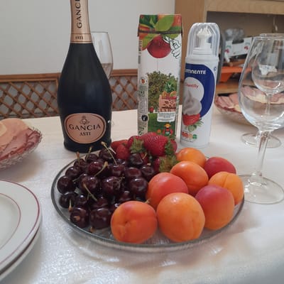 In Vila Verde, Portugal, a table is set with a spread of fruits and a bottle of Gancia Asti sparkling wine. A clear glass bowl overflows with dark cherries, their stems still attached, while another bowl holds sliced ham and cheese. Nearby, a carton of what appears to be juice or fruit puree and a can of Chantilly cream stand ready for use. Several wine glasses, one upright and another tipped, suggest a celebratory occasion. The scene is set against a backdrop of light-colored tablecloth and woven wicker furniture. The lighting is bright and natural, casting soft shadows across the table.