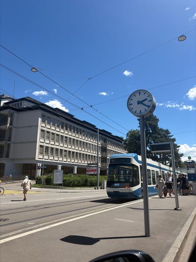 In Zurich, Switzerland, a bright, sunny day is captured on a street with a modern tram in motion. The tram, predominantly white with a blue stripe, has the number '6' displayed on its front and appears to be at a stop or slowing down. Several people are visible, some waiting at the tram stop, while others are crossing the street. One person in the foreground, seen from the back, is wearing a light-colored hat and shorts, walking across the tram tracks.

Dominating the scene is a large, circular clock on a pole, indicating the time to be approximately 10:09. Behind the tram and clock, a multi-story building with many windows lines the street. Overhead, wires crisscross the sky, suggesting an urban environment with a public transportation system. The sky is a vivid blue with scattered white clouds. The overall impression is one of a typical urban scene on a pleasant day in Zurich.