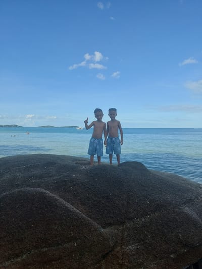 Two young boys, shirtless and wearing patterned swim shorts, stand on a large, dark rock at the edge of the water. The boy on the left raises his left hand, making a peace sign, while the boy on the right points forward with his right index finger. Both boys face the camera. Behind them, the ocean stretches to the horizon under a bright blue sky with scattered white clouds. In the distance, several islands are visible, along with a few people swimming and a boat on the water. The scene appears to be a beach or coastal area in Baan Chaweng Noi, Thailand.
