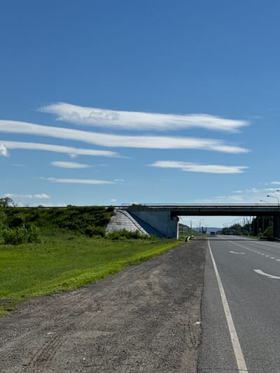 The image depicts a highway underpass on a bright, sunny day. The sky is a vibrant blue, populated by wispy, elongated white clouds. The underpass itself is a concrete structure with a metal railing on top. It spans over a paved road with clear white lane markings, including arrows indicating traffic direction. The road appears to be a multi-lane highway.

To the left of the road, a grassy embankment rises, covered in green vegetation. A portion of this embankment, near the underpass, is reinforced with concrete. Further to the left, some trees and bushes are visible. To the right of the road, streetlights are spaced along the roadside, and a line of trees forms a distant edge to the highway. In the distance, under the underpass, several vehicles, including what appears to be a truck, are driving on the highway.

The scene is set in an outdoor, rural or semi-rural environment, likely on the outskirts of a settlement. While the specific location in Krasnyy Yar, Russia, is mentioned, there are no visual cues within the image itself that definitively confirm this location. The image captures a common landscape feature of a modern highway infrastructure.