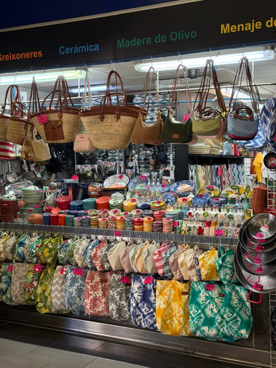 The image depicts a vibrant market stall in Palma de Mallorca, Spain, likely within a covered market such as Mercat de l'Olivar or Mercat de Santa Catalina, given the variety of goods. The stall is densely packed with an assortment of items for sale, primarily focusing on home goods and accessories.
In the upper section of the stall, a selection of straw and fabric tote bags are displayed, many with leather handles and some featuring price tags. Below these, rows of colorful ceramic plates, bowls, and decorative items are neatly stacked and arranged. The ceramics exhibit a variety of patterns, including floral motifs and geometric designs, with some items displaying handwritten price tags. Interspersed among the ceramics are smaller decorative bottles.
Further down, a collection of various fabric bags, many with bold floral and tie-dye patterns, hang from racks. These bags also have price tags indicating their cost. In the far right of the image, a stack of black metal frying pans and cooking utensils are visible, also with attached price tags.
The overall environment suggests a bustling retail setting, with bright lighting illuminating the products. The signs in the background, visible in the top portion of the image, include "Greixoneres," "Cerámica," "Madera de Olivo," and "Menaje de Oliva," which are Spanish and Catalan terms indicating that the stall specializes in pottery, olive wood products, and kitchenware. The merchandise suggests a focus on artisanal and decorative items, catering to both local residents and tourists. No people are visible in the frame.