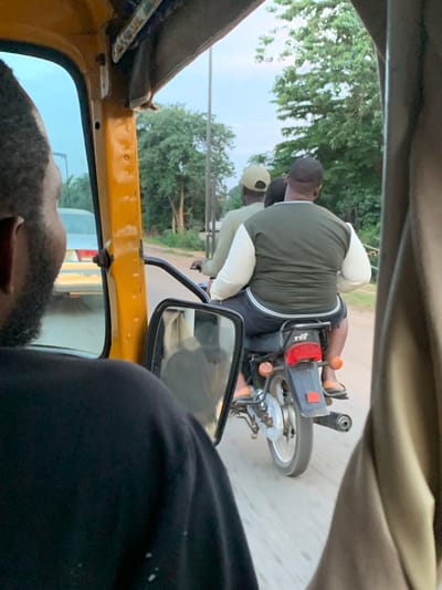 In the village of Galadimawa, Nigeria, two men are seen riding on a motorcycle on a paved road. The man in the rear, wearing a dark green and white shirt and dark shorts, is driving the motorcycle. The man in front of him, wearing a light-colored shirt and a light-colored cap, is the passenger. They are surrounded by greenery, with trees visible on the sides of the road. The image is taken from inside a yellow vehicle, likely a tuk-tuk, showing the back of the driver's head in the foreground, with a side mirror reflecting the road and surrounding environment. The sky appears overcast.