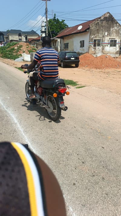 A man in a blue and orange striped t-shirt and dark shorts rides a red motorcycle down a paved road in Suleja, Nigeria. He is facing away from the camera, his left hand on the handlebars. His bare feet are on the footpegs. The motorcycle is a 2010s model with a black seat, chrome exhaust, and orange turn signals. The road has a white line marking on the left side and is bordered by a dirt shoulder. In the background, there are several buildings, including a house with a rusty metal roof and a black car parked in front of it. A pile of reddish-brown dirt is to the right of the house. Overhead, electrical wires are strung between utility poles. The sky is clear and blue. The foreground shows the blurred edge of a person's head or helmet, with black, yellow, and white stripes.