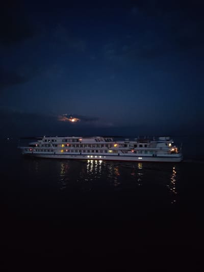 A wide, white, multi-decked river cruise ship is illuminated by its interior lights, sailing on dark water under a deep blue, cloudy night sky. The ship has several rows of windows, and its reflection is visible on the water's surface, broken by the ship's movement. A faint glow, possibly from the moon peeking through clouds, is visible in the upper left portion of the sky. The scene is dark, with only the lights from the ship and its reflection providing significant illumination. There are no people visible on the ship or in the surrounding environment. The image is a still photograph taken at night, depicting a tranquil but somewhat ominous maritime scene.