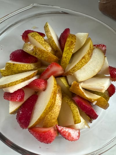 A close-up, overhead shot shows a clear glass bowl filled with sliced pears and strawberries. The fruit is arranged in a circular pattern, with the lighter colored pears alternating with the red strawberries. Some of the pear slices show green and yellow skin, while others are pure white flesh. The strawberries are cut into smaller, irregular pieces. The overall impression is of a fresh fruit salad. The scene is set in the city of Steklyannyi, Russia.
