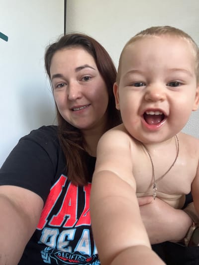 The image is a selfie of a woman holding a baby. The woman is on the left, and the baby is on the right, held in her arms and partially obscuring her. The woman has long brown hair and is wearing a black t-shirt with red and white lettering. The baby has light-colored hair and is facing the camera with an open mouth, appearing to be laughing or shouting. The background is a plain, lightly textured wall. The scene appears to be indoors, likely in a residential setting. There is no specific visual information to place this image in Chelyabinsk, Russia.