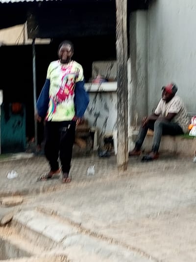 In Arawa, Nigeria, a blurry outdoor scene captures two individuals. In the foreground, a person stands facing slightly left, wearing a colorful t-shirt with a pattern of pink, yellow, green, and blue, and dark pants. Their arms are extended outward, covered by a dark blue garment. They are wearing sandals. To the right and further back, another person is seated on a bench, facing left. They wear a light-colored t-shirt and dark pants, with a red and black baseball cap. The environment appears to be an unpaved area with some debris on the ground, and a structure with a dark roof is visible in the background. The lighting suggests it is daytime.