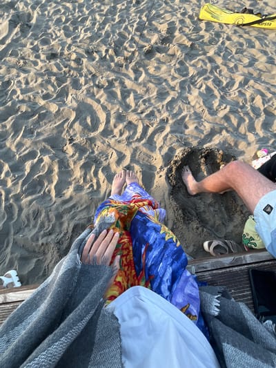 The image shows a top-down view of a sandy beach in Pistula, Montenegro. Two people are present, partially visible. One individual's legs are extended, with their right foot in a dug-out sand hole. This person is wearing light-colored swim shorts. The other individual's legs are bent, covered by a colorful patterned fabric and a grey blanket. Their hands are visible, one with tattoos.
The beach is covered in rippled sand, suggesting wind or water movement. Scattered on the sand are a yellow Kipsta beach volleyball bag, a pair of flip-flops, and a white sandal. A wooden structure, possibly part of a bench or boardwalk, runs horizontally across the lower portion of the image. A black smartphone is placed on this structure to the right. The lighting suggests it is late afternoon or early evening.