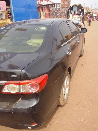 The image captures a black Toyota Corolla LE sedan parked facing away from the viewer, angled slightly left, on an unpaved, reddish-brown dirt road in Ankpa, Nigeria. The vehicle's exterior exhibits an accumulation of dust and dirt, particularly on the trunk lid and rear bumper. The rear bumper shows multiple scratches and areas of paint loss, notably on the lower left corner and across the bottom edge. The right taillight assembly is visible and intact. The rear window is tinted, displaying several light-colored stickers and reflections, with a light-colored object visible on the interior rear shelf. The right side of the vehicle shows four doors, each with a chrome handle, and the right rear wheel is visible, also coated in dust.
In the background to the left of the sedan, a prominent bright blue metal gate or shutter is visible. Adjacent to it are stacks of merchandise, including what appear to be mattresses or cushions, one featuring a graphic of a smiling woman's face. Further back, red crates and other stacked industrial materials, possibly tires, are present.
To the right of the sedan, the unpaved road extends into the distance, revealing an active outdoor market or street scene. Multiple individuals are visible, indicating pedestrian traffic. One person is pushing a loaded two-wheeled cart topped with a makeshift canopy from which clothing items are suspended. Other indistinct figures and vehicles can be seen further down the street. The environment suggests a busy commercial area under daylight conditions.