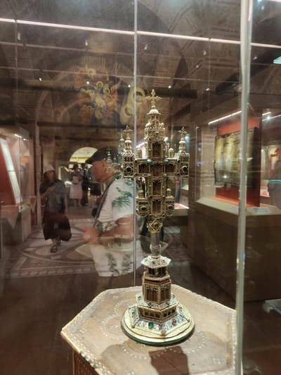 A golden reliquary cross, adorned with small gemstones and intricate architectural details, stands on a decorative wooden pedestal within a museum display case. Reflections in the glass reveal a museum interior, including other exhibits and visitors. In the background, a wall painting depicts figures, possibly religious in nature. A man wearing a patterned shirt and a cap, and a woman with a hat and a long skirt, are visible among the museum visitors, their figures blurred by reflections. The floor of the museum is visible, with patterned tiles and a dark brown polished surface. The overall scene suggests a cultural exhibition in a public institution. The image was taken in the city of Kalambaka, Greece.
