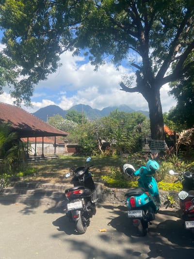 An eye-level, medium shot captures a sun-drenched scene featuring parked motorbikes in front of a traditional Indonesian building with a red tile roof, set against a backdrop of lush greenery and distant mountains. Three motorbikes are visible: a black Yamaha NMAX on the left with license plate DK 2786 UBJ, a teal scooter in the center with license plate DK 5894 FDD, and a partial view of a red motorbike on the right with license plate DK 2133. Two helmets, one white and one teal, are placed on the handlebars of the teal scooter. The building, likely a temple or guesthouse, has open-air verandas with wooden posts. Dense foliage, including palm trees and flowering bushes, surrounds the property. The weathered mountains in the background are partially obscured by a light haze. The sky above is a mix of blue with scattered white clouds. The lighting suggests it is midday. The scene is located in Gerokgak, Indonesia.