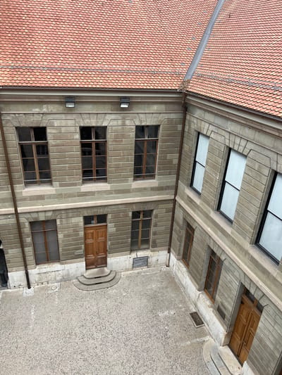This image captures a high-angle view looking down into an inner courtyard of a building in Genève, Switzerland. The scene is static and devoid of people, illuminated by soft, diffused daylight.
A prominent feature at what appears to be the top edge of the courtyard is a section of a red and orange terracotta tiled roof. A grey metal gutter runs along its lower edge, containing some natural debris like leaves or a small nest.
To the left, a multi-story stone facade forms one side of the courtyard. It is constructed from light grey-beige rectangular blocks, featuring several rows of recessed rectangular windows with dark frames, likely wooden. The masonry exhibits a structured pattern, conveying a sense of age and solidity. Multiple wall-mounted light fixtures and a vertical downspout are visible on this facade.
The courtyard floor occupies the lower and right parts of the image, paved with a light-colored, speckled material, possibly concrete with fine gravel. Two sets of gently curved stone steps lead up to recessed entrances, each fitted with traditional wooden double doors.
Above the courtyard, integrated into the upper structure on the right, are several rectangular skylights and smaller windows with wooden frames, contributing to the diffused natural light within the space.
The architectural style suggests a historical European building, possibly institutional or a large residential complex, consistent with its location. The even lighting implies an overcast day or indirect illumination within the enclosed area. No visible text is present.