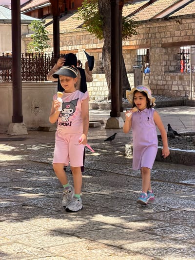 The image shows two young girls enjoying ice cream cones while walking on a cobblestone path. The girl in the foreground, wearing a pink t-shirt with a Kuromi character and pink shorts, is holding a cone in her right hand. She is also wearing a black and white striped hat and silver sneakers with green socks. The girl in the background, wearing a light purple dress and a straw hat, is also holding an ice cream cone. She has her tongue sticking out and appears to be enjoying her treat. They are walking in a sunny outdoor environment, possibly a street or courtyard, with stone buildings and a wooden railing visible. Pigeons are present on the ground around the girls. The overall mood seems cheerful and summery.