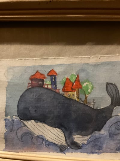 This is a close-up watercolor painting depicting a whimsical scene. The central subject is a large, dark blue whale that appears to be emerging from blue water with white waves. Perched on the whale's back is a small village scene with red-roofed houses and green trees. The houses are rendered in a simplistic style, with some appearing to be multi-story with windows.
The painting has a soft, artistic feel due to the watercolor medium. The background is a light blue, suggesting a sky or calm water. The overall mood is surreal and imaginative, perhaps evoking a sense of wonder or a dreamlike state. There are no visible people. The painting is framed by a light wooden border. The artwork's style suggests it could be a folk art or illustrative piece. No specific time of day, weather, or clear location cues are evident beyond the village and sea elements. The location context of Sergeevo, Russia, does not provide direct visual cues within the painting itself. There is no visible text in the image.