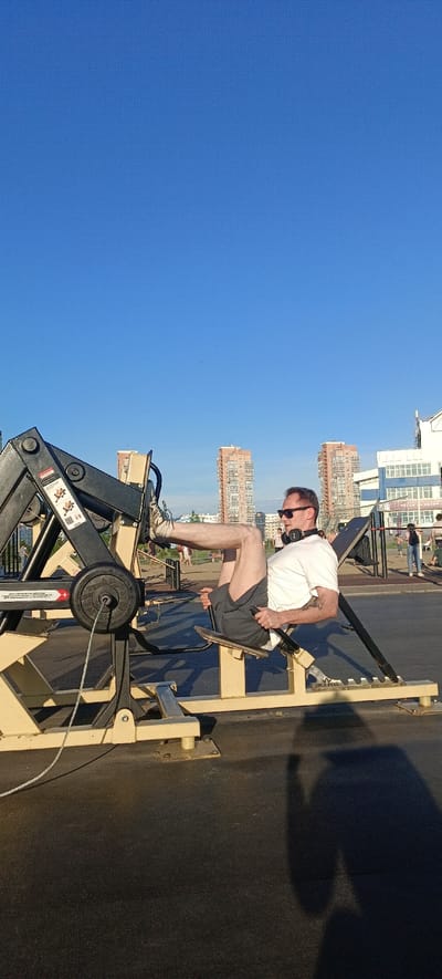 A man is exercising on a leg press machine outdoors. He is wearing a white t-shirt, grey shorts, sunglasses, and headphones around his neck. His legs are extended, pressing against the machine's platform. The scene is an outdoor gym with other exercise equipment and people in the background. In the distance, two tall, peach-colored apartment buildings are visible, with a clear blue sky above. The time of day appears to be late afternoon or early evening, as indicated by the long shadow cast by the man and the lighting. The setting suggests a public park or recreational area, possibly in an urban environment given the buildings. There is text visible on the leg press machine, including "IRON KING" and some warning symbols with instructional diagrams. The overall mood seems to be one of fitness and activity in a pleasant outdoor setting.