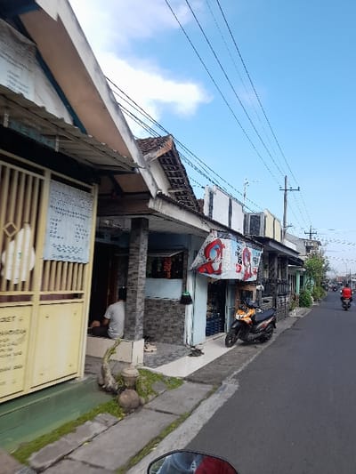 The scene is a street in Kecamatan Tanggulangin, Indonesia, with coordinates -7.505 latitude and 112.718 longitude. There are several buildings on the street, including a shop with a sign that says "99".  The street is paved and there are power lines overhead. There is a motorcycle parked on the street. There is a man sitting on the ground in front of a building. There is vegetation growing on the ground and on the side of the street. The sky is blue with some clouds.