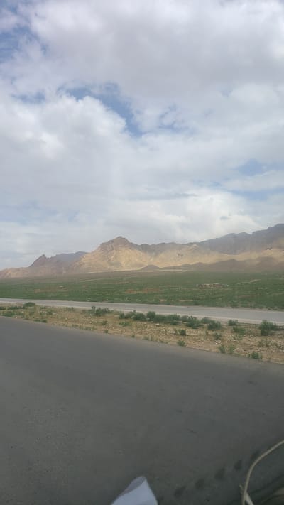 The image displays a vast outdoor landscape under a partially cloudy sky. The sky, occupying the left side of the frame, is filled with large, white and light grey clouds, with patches of clear blue sky visible in between.
In the mid-ground, a long, rugged range of barren mountains stretches across the horizon. These mountains are predominantly light brown and tan, showcasing a dry, rocky terrain devoid of significant vegetation, characteristic of an arid or semi-arid region.
The foreground is dominated by a dark grey, paved road or highway that runs diagonally from the bottom right towards the center. Adjacent to the left side of this road is a prominent strip of vibrant green land, which appears to be cultivated fields or lush pastures, contrasting sharply with the parched, sandy earth surrounding it and the distant mountains. Sparse, low-lying green vegetation is scattered along the edges of the road and on the dry ground. On the far right edge, a small white object, possibly a piece of cloth or paper, and some thin, dark lines, possibly cables, are partially visible. The perspective suggests the photograph was taken from a vehicle.
The scene depicts a wide-open, natural environment in a mountainous area. The lighting indicates it is daytime, likely late morning or afternoon, given the diffused light filtering through the clouds. No people or specific activities are visible. The landscape is consistent with the geographical characteristics of Khanai Baba, Pakistan. No visible text is present in the image.