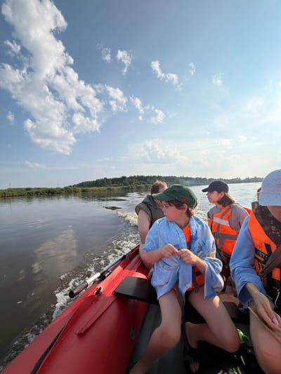 The image depicts a bright, sunny day with scattered clouds in the sky, as viewed from the perspective of someone on a red inflatable boat moving through a body of water. The boat is carrying several individuals, including children and adults, all wearing bright orange life vests. In the foreground, a young person in a green baseball cap and a blue patterned wrap over an orange life vest is seated. To their right, another person wearing a dark baseball cap and an orange life vest is visible. Further back, a person with darker hair and an orange life vest is also in the boat. The water is calm with a visible wake trailing behind the boat, indicating movement. The background features a lush green shoreline with a dense forest and some vegetation. The scene appears to be a recreational outing on a river, likely in or near the village of Kalino, Russia, given the context provided. The overall impression is one of a leisurely boat trip on a summer day.