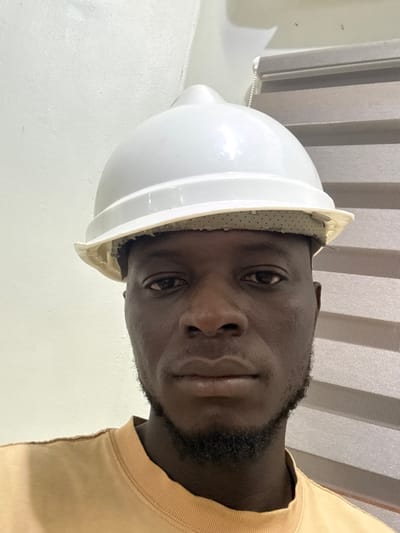 An adult male, with dark skin, a short beard, and mustache, is depicted from the chest up, looking directly forward. He wears a white hard hat, positioned on his head with a visible, light-colored, perforated inner lining showing beneath the brim. He is dressed in a light brown, long-sleeved shirt. The background, an indoor setting within the city of Lafia, Nigeria, features a plain white wall on the left side of the frame and vertical window blinds with alternating light and dark grey horizontal stripes on the right. A white pull cord for the blinds is visible.